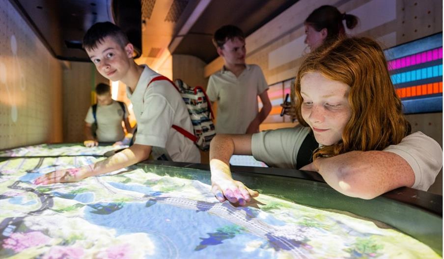 Several school-aged children interact with a large, illuminated interactive display table that shows a colorful map or simulation, inside a modern, di