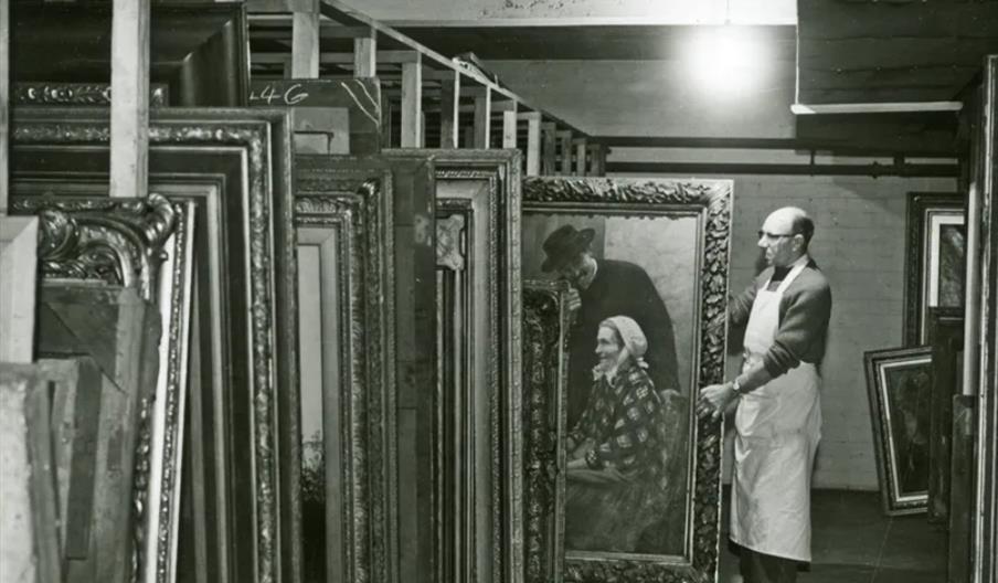 A man wearing an apron stands in a storage room at the Ferens Art Gallery, holding a large framed painting among rows of other stored artworks. The sp