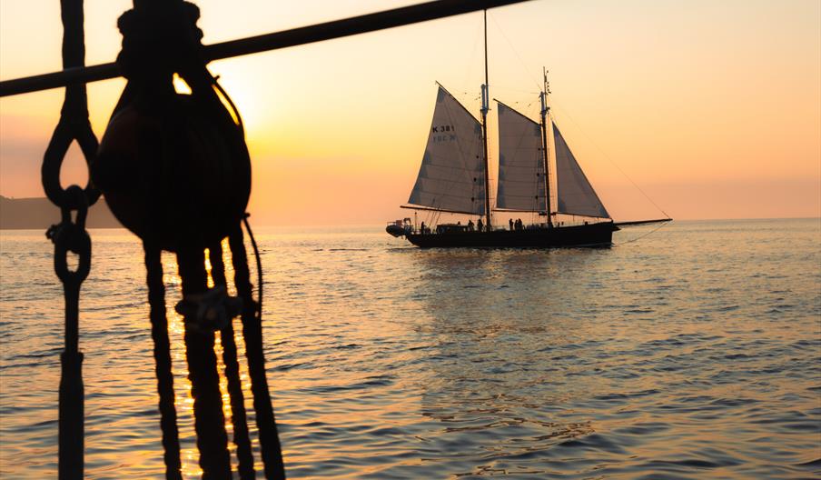 A sailing ship with two large sails glides across calm water at sunset, with the sky glowing orange and the silhouette of rigging in the foreground.