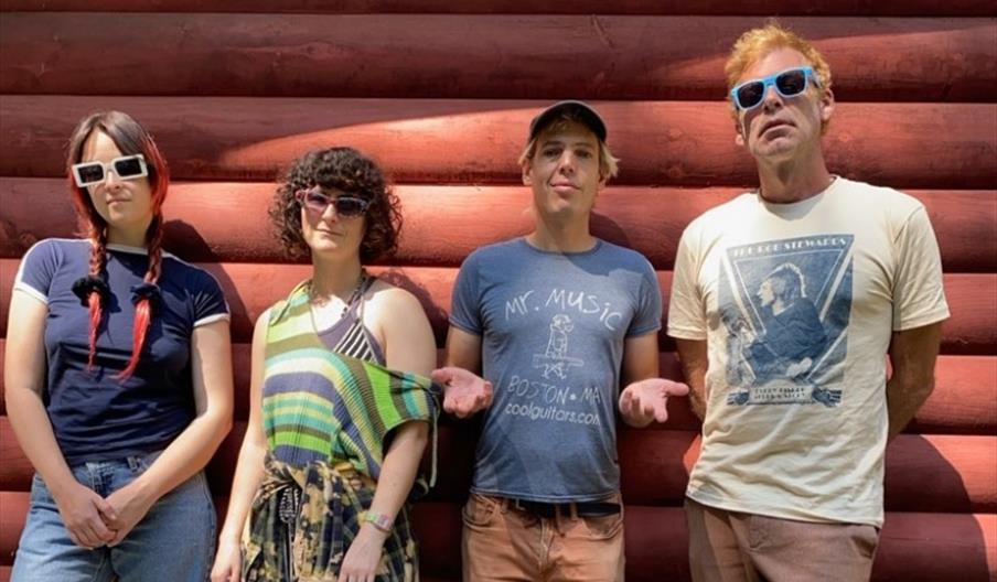 Four people stand side by side in front of a red log wall, each wearing sunglasses and casual clothing, with sunlight casting shadows behind them.