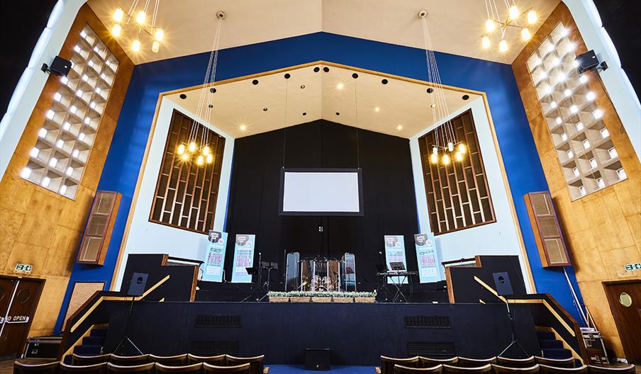 A wide view of a modern auditorium or church sanctuary featuring a raised stage with musical instruments, banners, and a large blank screen at the cen