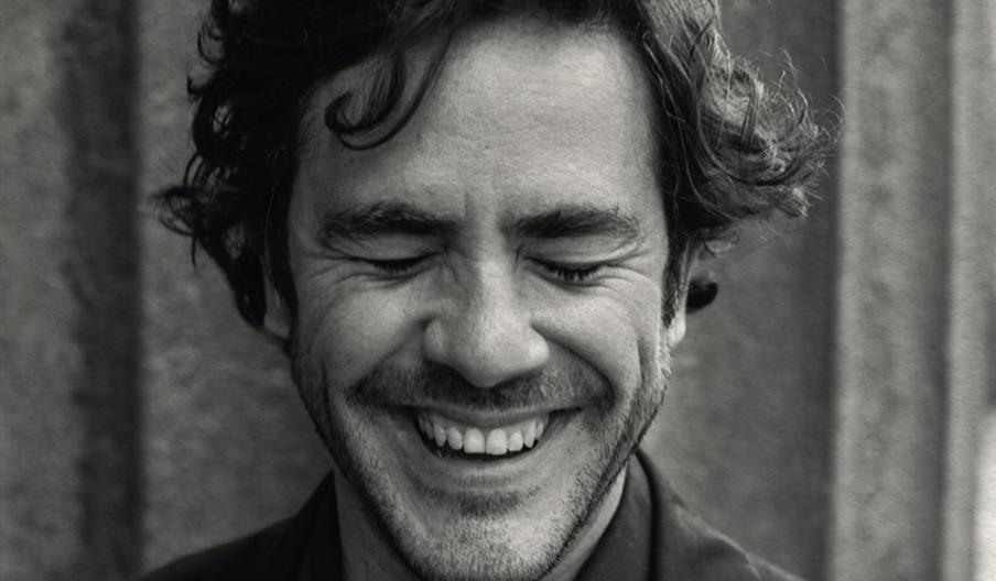 Jack Savoretti smiles broadly with his eyes closed, appearing joyful in this black-and-white close-up portrait.