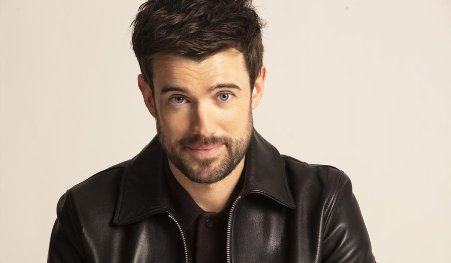 Jack Whitehall comedian, stirring into the camera, wearing a black polo shirt and black leather jacket.