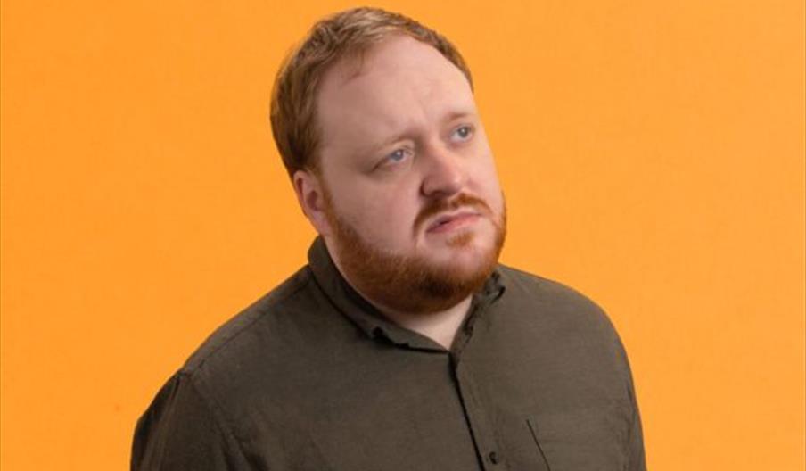 A man with short reddish hair and a beard wearing a dark shirt, comedian Jamie Hutchinson stands against a solid orange background, looking slightly u