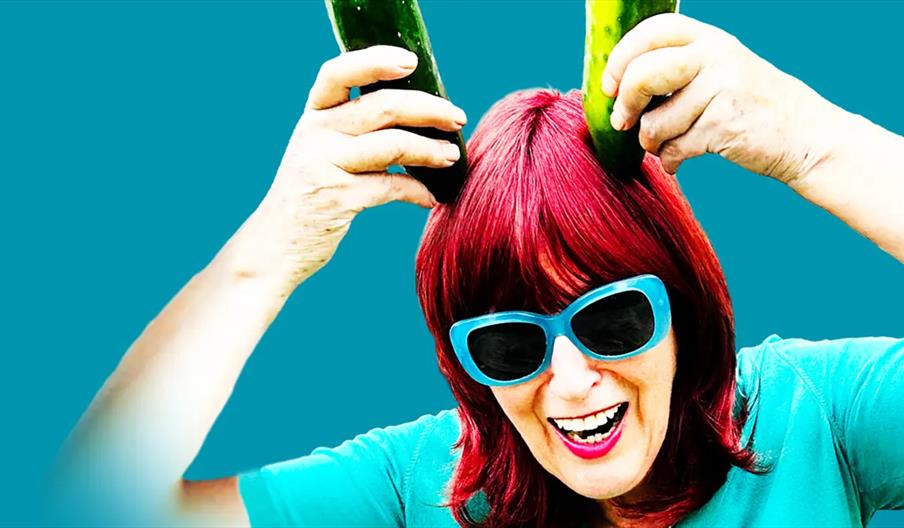A smiling person with red hair and blue sunglasses playfully holds two cucumbers on top of their head like horns against a blue background.