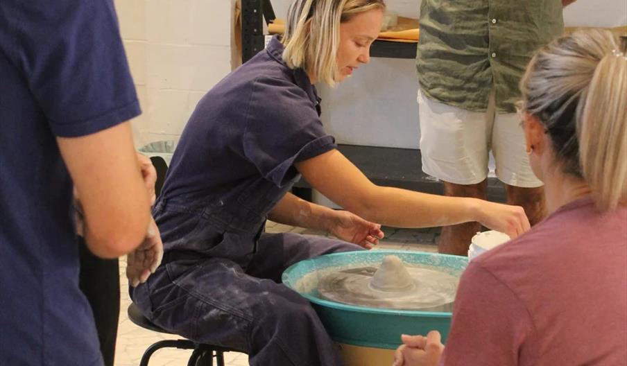 A woman sits at a pottery wheel shaping clay while three people watch her demonstration in a studio with shelves and supplies in the background.