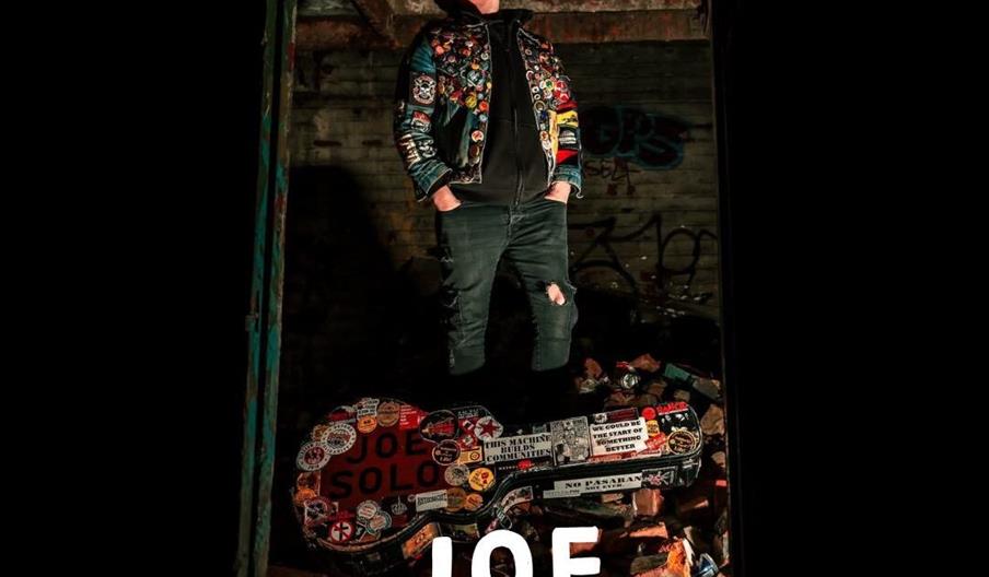 A person wearing a colourful patch-covered jacket and ripped jeans stands among rubble in a dimly lit, graffiti-marked urban space, looking upward. In