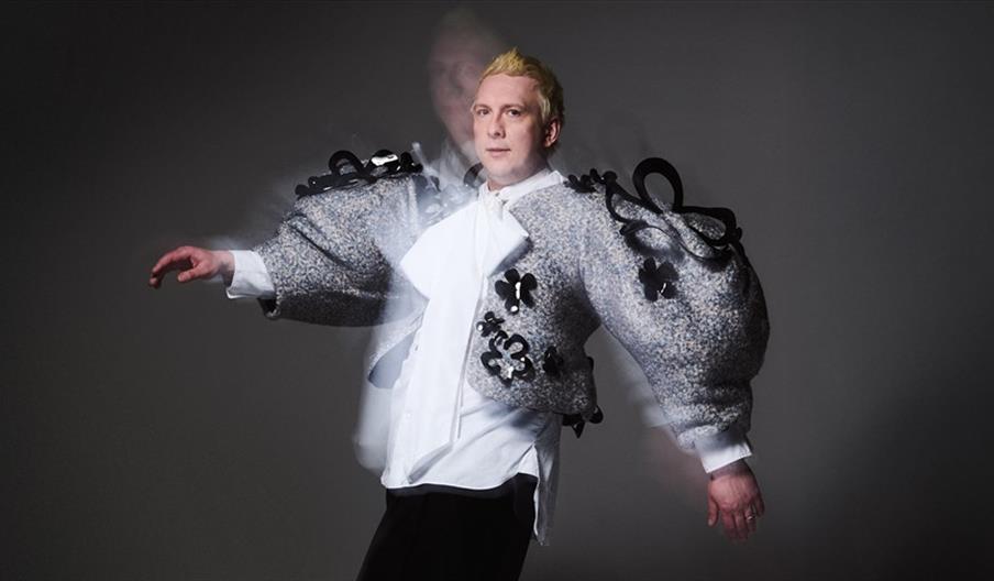 A person with short blond hair wears an oversized, textured jacket with dramatic puffy sleeves and black decorative accents over a white shirt, standi