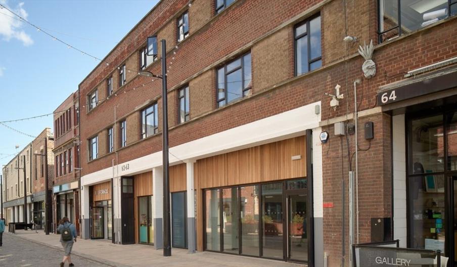 A street view of a row of brick buildings on Humber Street with large windows and shopfronts, including a gallery at number 64. Several people walk al