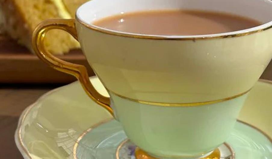 A pastel green and gold teacup filled with tea sits on a matching saucer, with slices of yellow cake in the background on a wooden table.