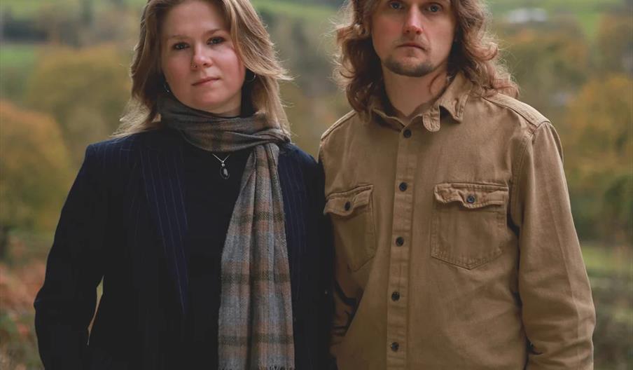 Two people stand outdoors in front of a blurred green landscape. The person on the left has shoulder-length light hair, is wearing a dark jacket, and