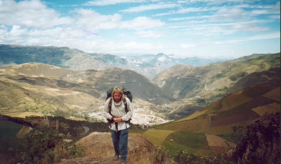 A person with a backpack hikes up a dirt trail on a mountain, with a wide view of valleys, patchwork fields, and distant mountains under a partly clou