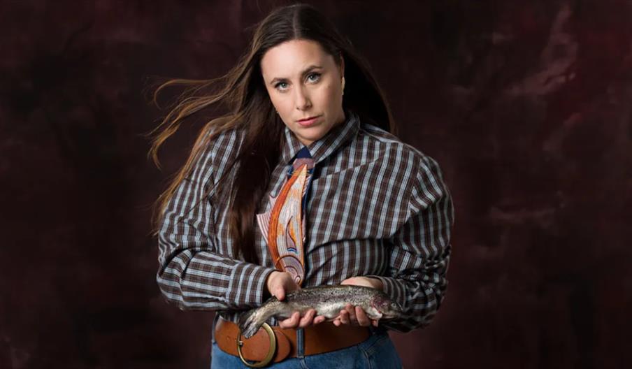 A woman with long brown hair, wearing a plaid shirt and colorful tie, stands against a dark background holding a fish in both hands and looking direct