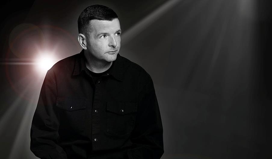 Kevin Bridges is wearing a dark button-up shirt sits against a dark background, with a bright light shining from behind him and dramatic light rays vi