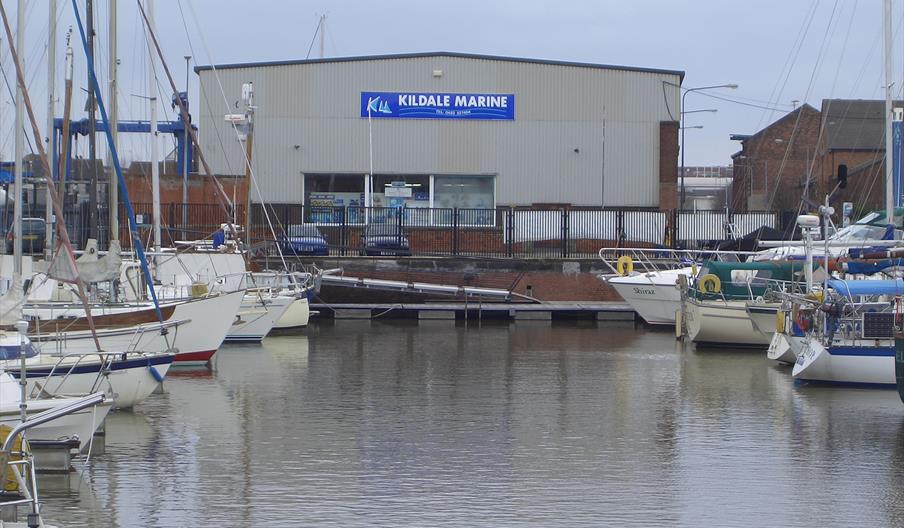 Several sailboats are docked in a marina, with a large building labeled "Kildale Marine" in the background. The water is calm and the sky is overcast.