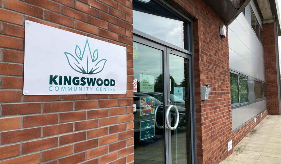 The entrance to a modern brick building with a glass door, displaying a sign on the wall that reads "Kingswood Community Centre" with a leaf logo abov
