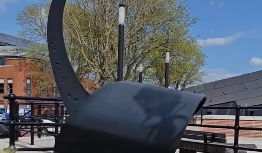 A large black metal sculpture resembling part of a ship’s anchor stands beside a waterside walkway, framed by spring flowers in the foreground. Trees