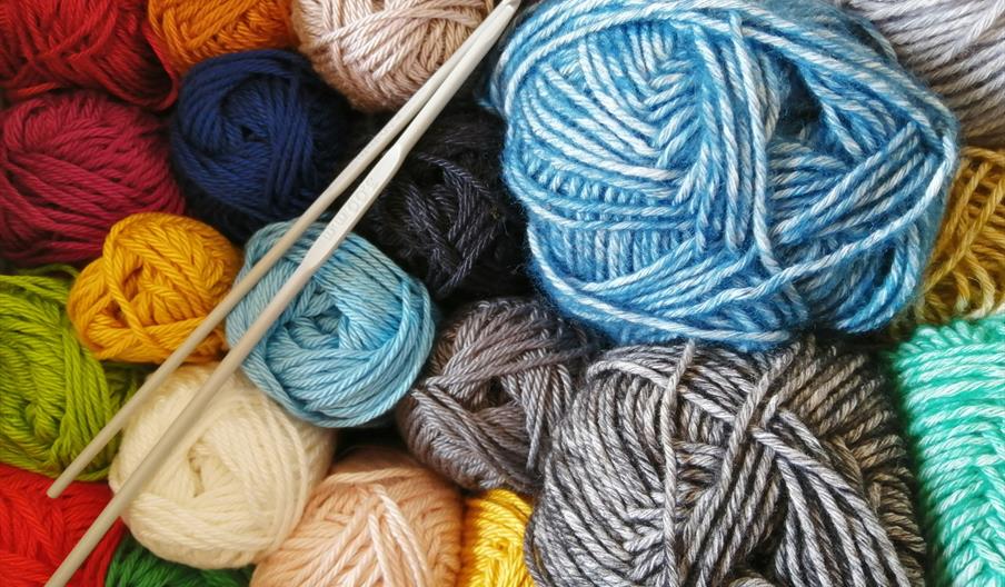Several balls of yarn in various colors, including blue, red, yellow, green, and gray, are closely arranged with two knitting needles resting on top.