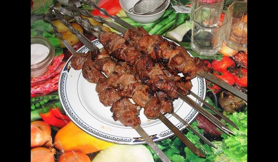 Several metal skewers with grilled meat are arranged on a decorative plate placed on a colourful tablecloth featuring images of vegetables. There are