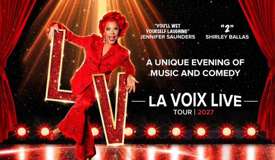 A performer with bright red hair and a sequined red suit poses energetically on stage, holding giant glittering red letters "L" and "V." The stage is