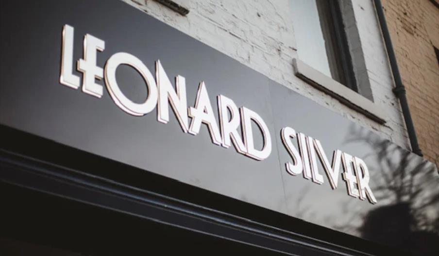 A black storefront sign with white, bold letters that read "LEONARD SILVER" mounted on a building with a white brick wall and window above.