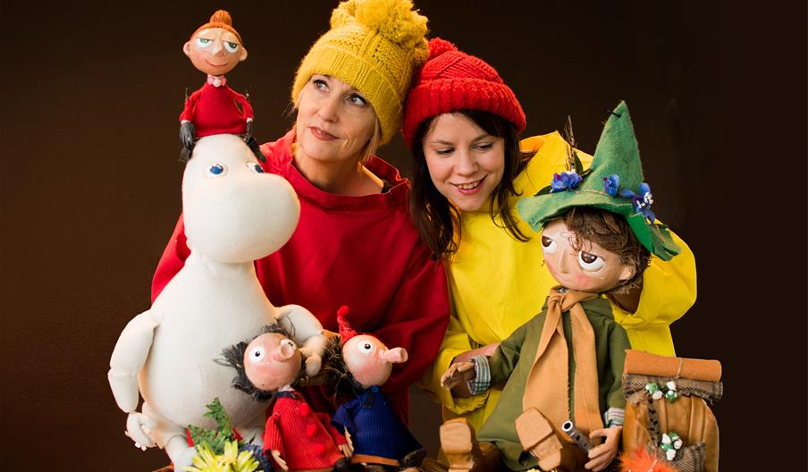 Two women, one wearing a yellow hat and red shirt and the other in a red hat and yellow shirt, are smiling while surrounded by Moomin characters as pu