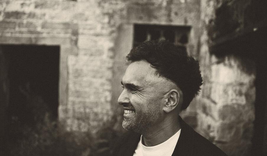 A man with short, dark hair and a trimmed beard smiles while standing outdoors in front of an old stone building. The image is in black and white, giv