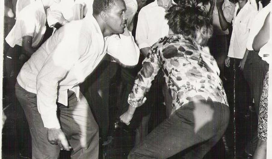 Black and white photo of a lively dance scene from the mid-20th century, showing a group of young people dancing energetically, with a man in a light