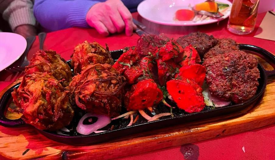 A sizzling platter of assorted Indian appetizers, including reddish tandoori chicken pieces, brown onion bhajis, and kebabs, sits on a wooden board at