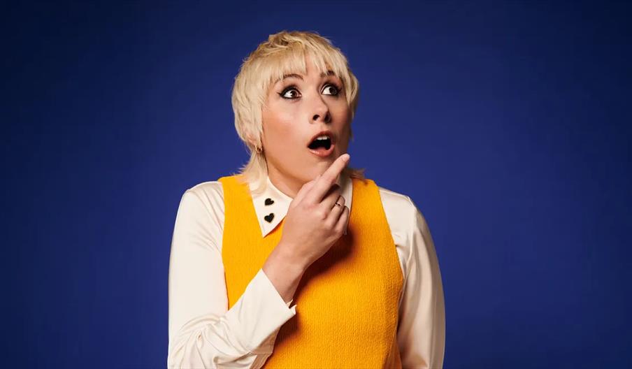 Maisie Adam with short blond hair wearing a yellow sleeveless top over a white shirt stands in front of a blue background, looking surprised with thei