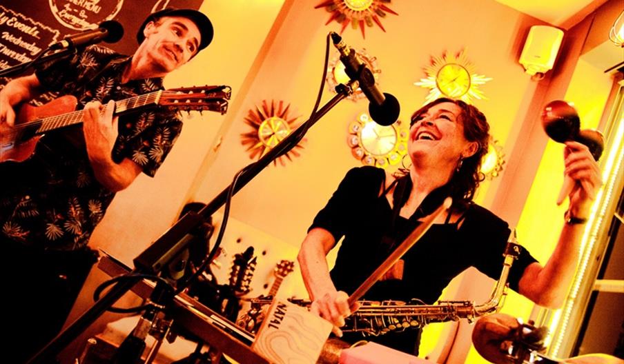 Two musicians perform enthusiastically indoors; one plays a guitar and sings at a microphone, while the other smiles, holding maracas and a saxophone.