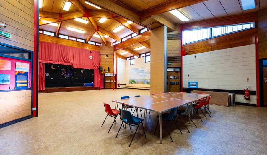 Interior of Maurice Rawling Centre. A spacious multipurpose hall with a high wooden ceiling, a small stage with red curtains at one end, and a cluster