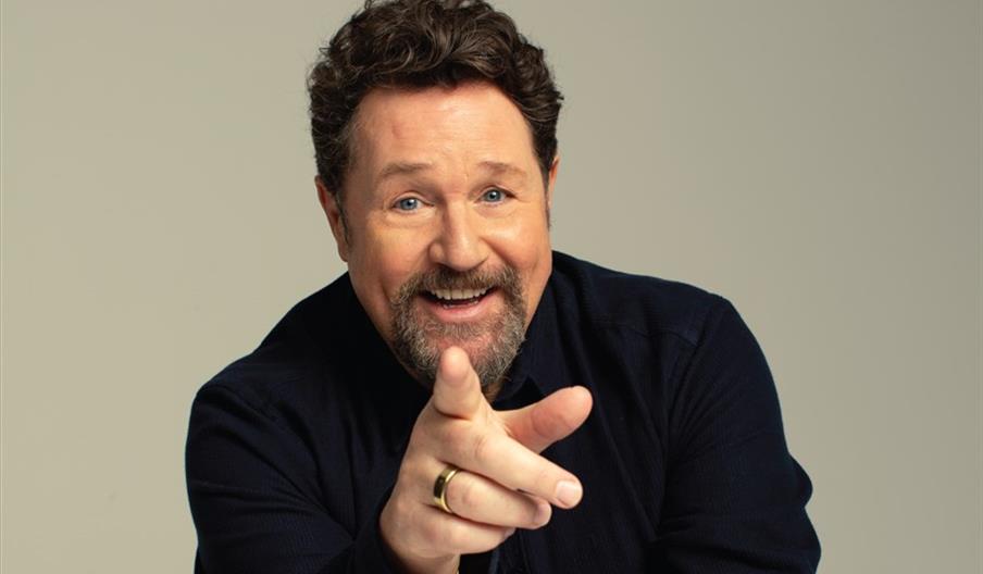 Michael Ball is smiling and pointing toward the camera, wearing a dark shirt against a plain, light background.