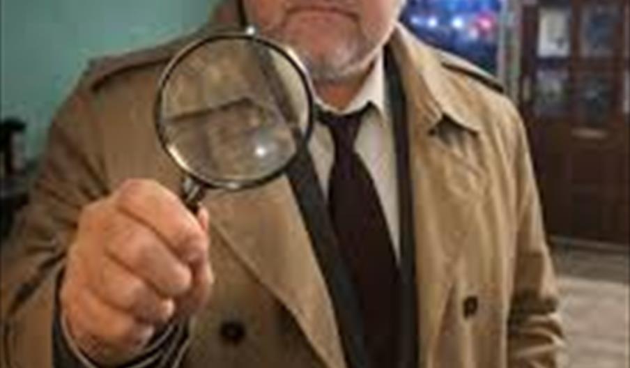 A man dressed as a detective in a trench coat and fedora holds a magnifying glass up toward the camera, looking serious.