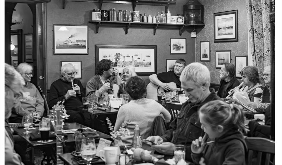 A black and white photo of a lively pub scene shows people gathered around tables, some playing musical instruments like guitars and a tin whistle, wh