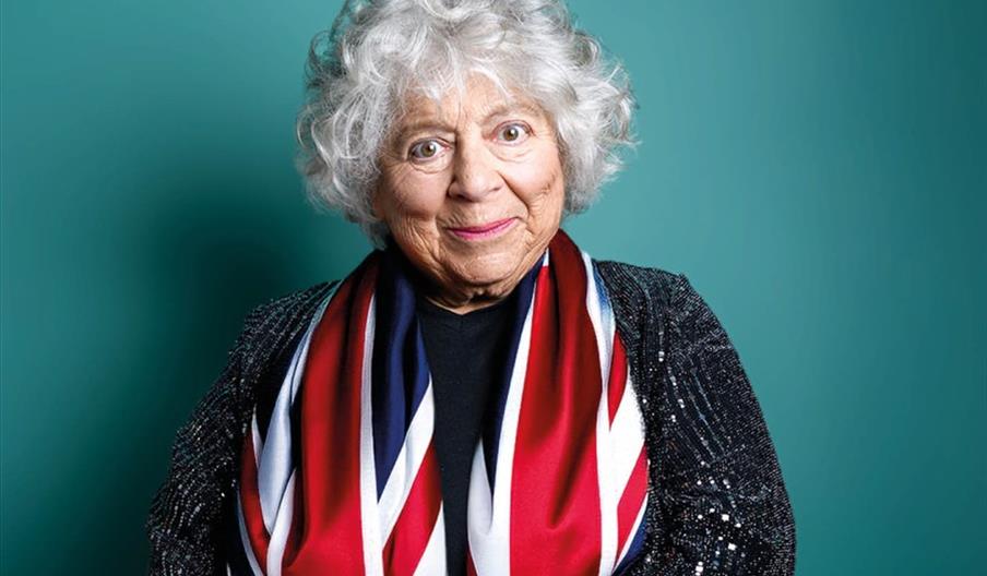 Miriam Margoyles smiles at the camera, wearing a sparkly black jacket and a red, white, and blue scarf, standing in front of a teal background. She's