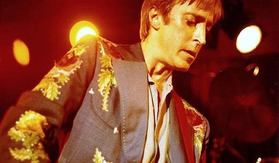 A musician wearing a colorful jacket with floral embroidery is playing an electric guitar on stage, illuminated by warm, orange stage lights in the ba