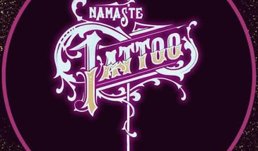 A circular, ornate logo with the words "Namaste Tattoo" in stylized, decorative lettering, featuring intricate swirls and a pointed vertical design, s