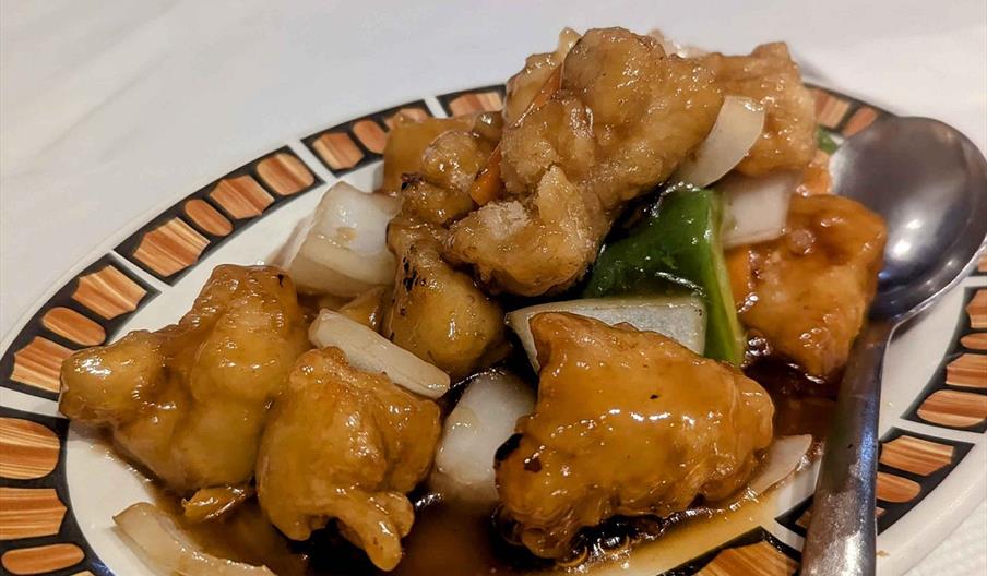A plate of sweet and sour chicken featuring golden-brown battered chicken pieces, sautéed onions, and green peppers served in a glossy brown sauce, wi