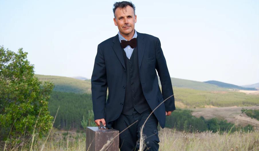 A man wearing a dark suit and bow tie walks through a grassy field carrying a suitcase, with rolling green hills and open sky in the background.