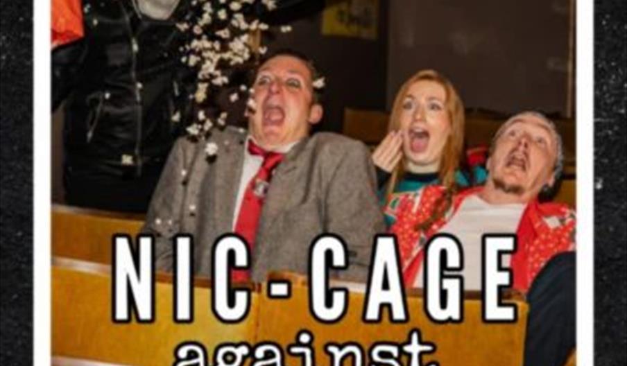 A person in a flaming orange and yellow mask and black leather jacket is jumping over theater seats, throwing popcorn at three shocked and scared peop