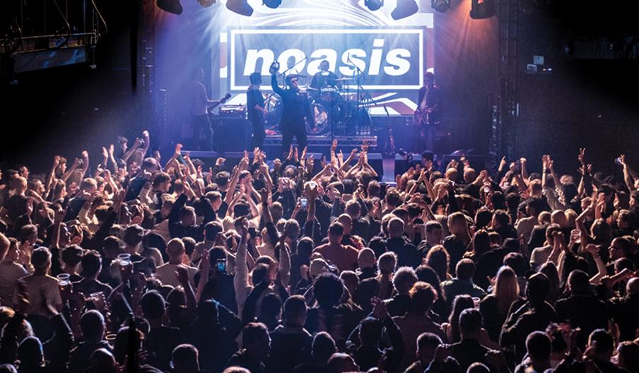 A large, energetic crowd faces a stage where a band performs under bright lights, with the word "noasis" displayed on a screen behind them. The scene