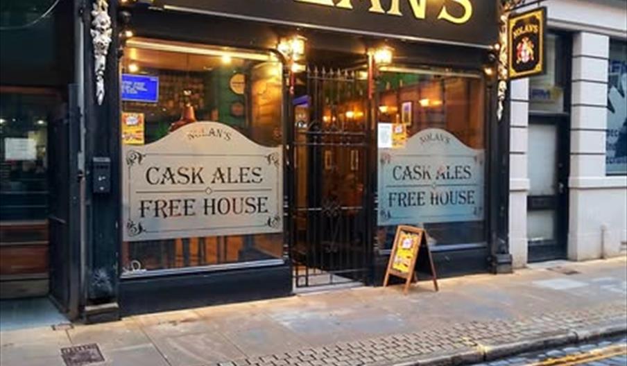 A traditional pub with a black façade and gold sign reading "NOLAN'S" above the entrance, flanked by large windows advertising "Cask Ales" and "Free H