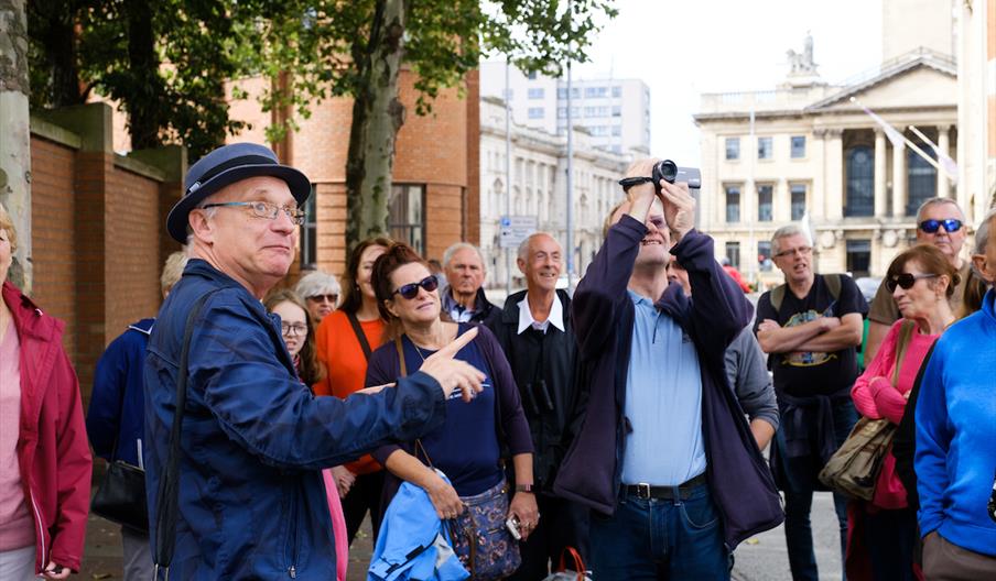 A group of people stands outdoors in a city; a man in a blue jacket and hat gestures and speaks while another man films with a small video camera, and