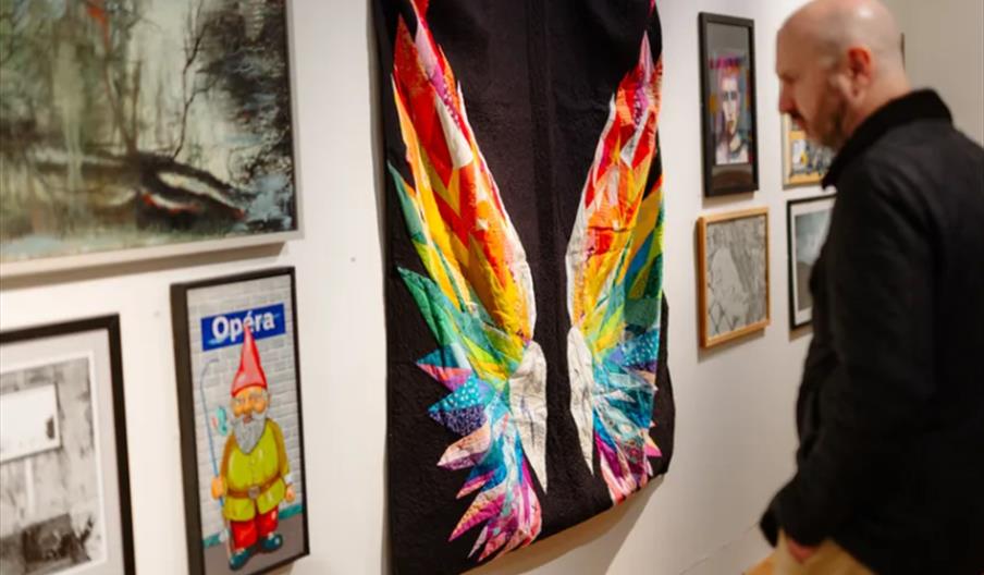 A person stands in a gallery looking at a quilted wall hanging featuring large, colorful patchwork wings on a black background, surrounded by various