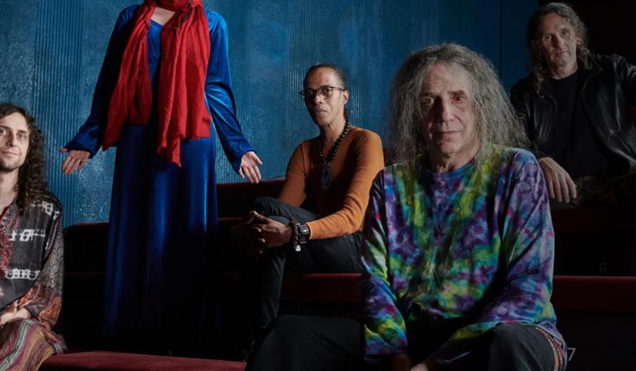 Group Ozric Tentacles, five people sit and stand on red theater seats against a blue wall, with one person in a blue dress and red scarf standing whil
