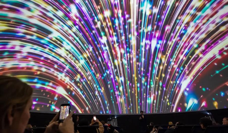 People seated in a planetarium watch a large, colorful light display resembling an explosion or fireworks projected on a domed screen, with some audie
