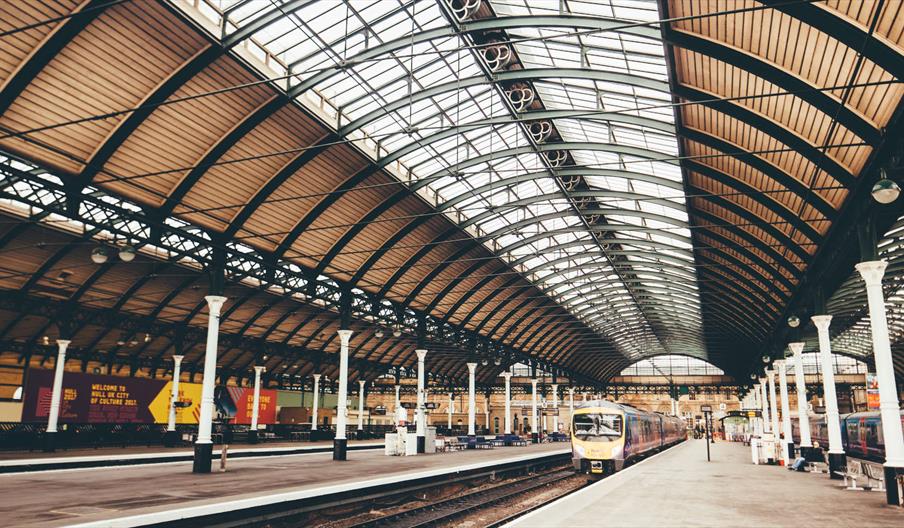 A spacious indoor train station with a high arched glass and metal roof, multiple platforms, and a train waiting on one of the tracks. The station is