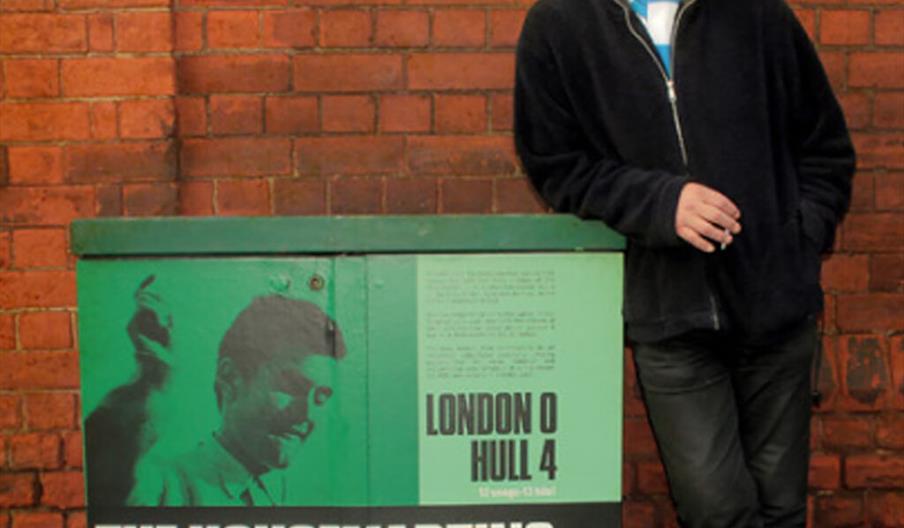 An older man wearing a black cap and jacket leans against a green utility box in front of a brick wall. The box features a large image, bold text read