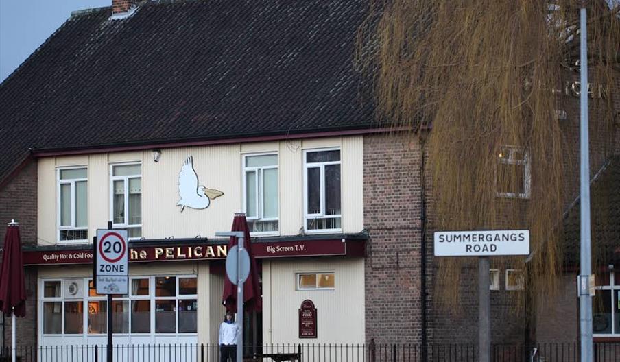 A two-story pub named "The Pelican" with a brown tiled roof, cream and white exterior, and red umbrellas shading outdoor seating in front. A large pel
